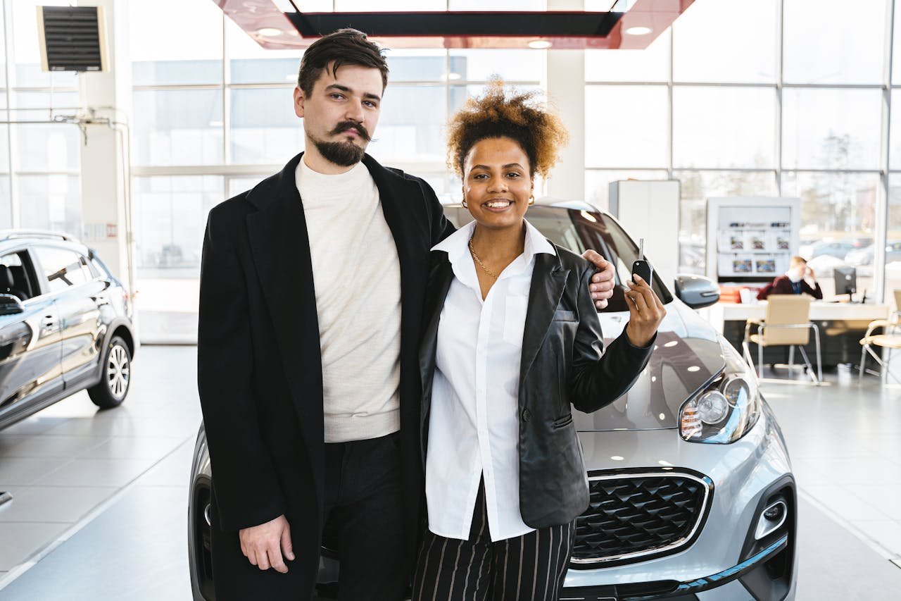Interracial couple celebrates buying a new car at a dealership, holding keys and smiling.